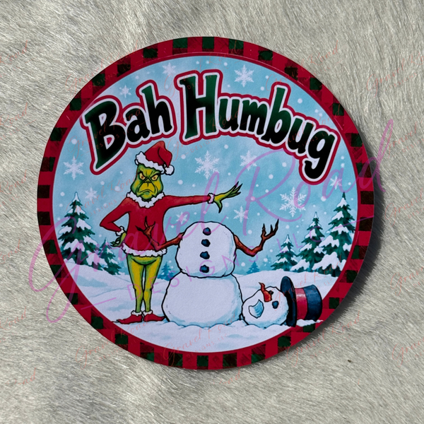 A circular cardstock featuring a Christmas-themed G illustration with the text 'Bah Humbug' on a snowy background, placed on a gray textured surface.