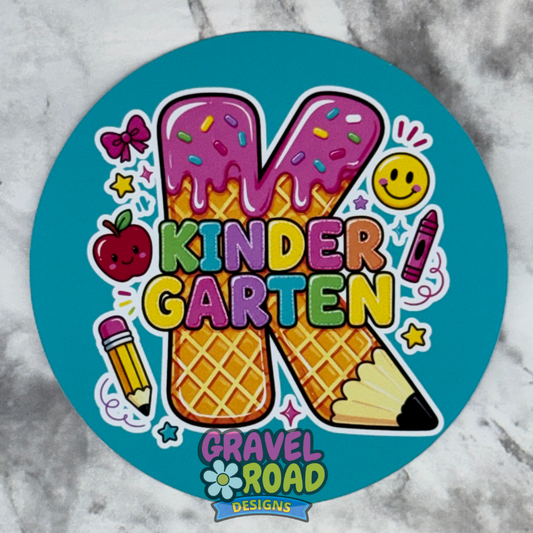 A round cardstock with a colorful kindergarten theme, featuring a large letter K made of waffle cones and ice cream cones, with the words 'KINDER GARTEN' and the logo 'GRAVEL ROAD DESIGNS' on a turquoise background, shown on a gray marble surface.