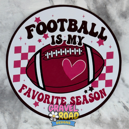 A circular cardstock design featuring a football with a red heart in the center and the text 'FOOTBALL IS MY FAVORITE SEASON' in dark brown and red, with red checkerboard accents on a white circle. A 'Gravel Road Designs' logo is visible at the bottom.