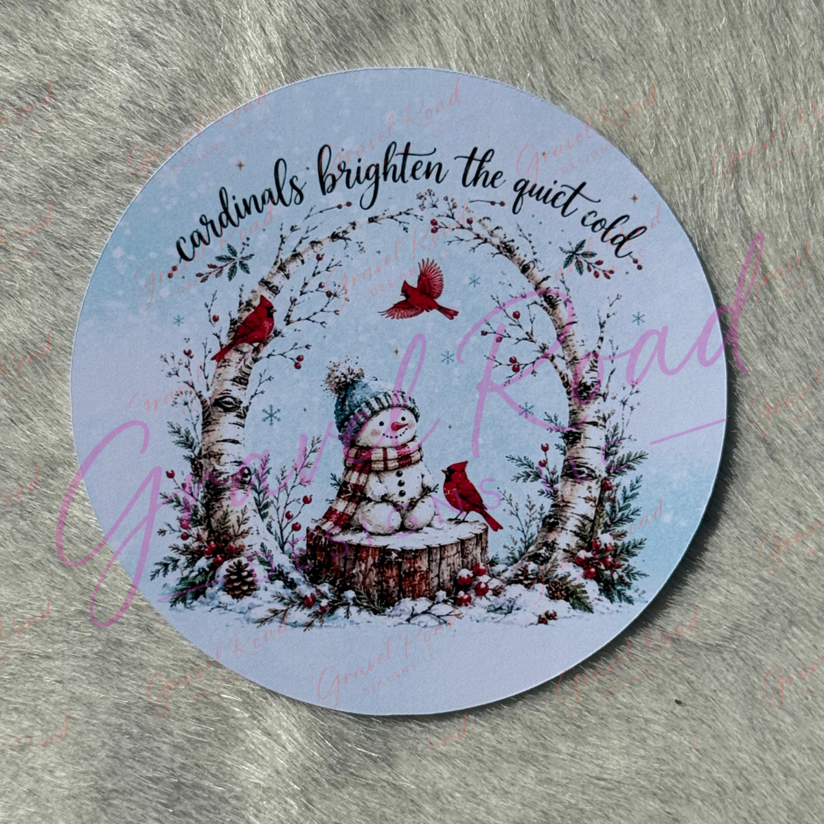 A round printed cardstock featuring a winter illustration with a snowman, birch tree, red cardinals, and the phrase 'cardinals brighten the quiet cold' on a pale blue background, displayed on a gray textured surface.