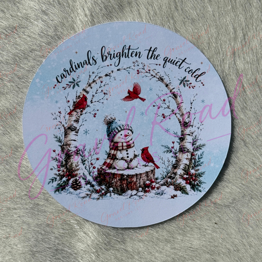 A round printed cardstock featuring a winter illustration with a snowman, birch tree, red cardinals, and the phrase 'cardinals brighten the quiet cold' on a pale blue background, displayed on a gray textured surface.