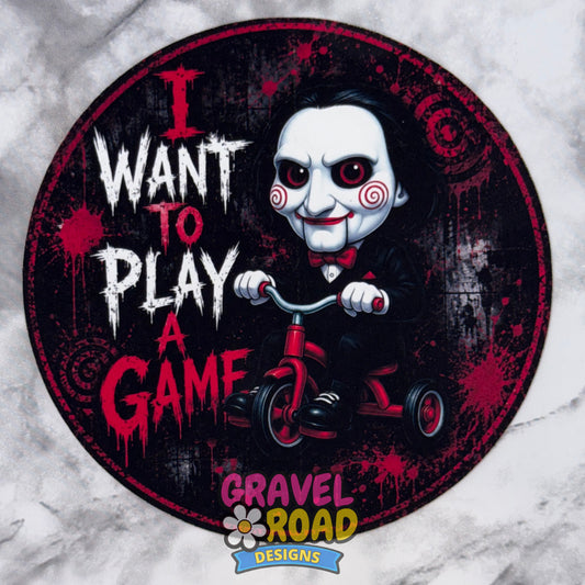 A circular cardstock design featuring a cartoon-style evil child with a white face, red eyes, and a black suit riding a red tricycle. Large distressed text reads 'I WANT TO PLAY A GAME' in white and red. Gravel Road Designs logo at the bottom. On a grey marble backdrop.