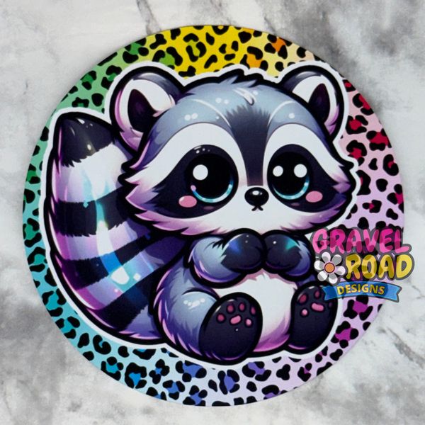 A circular cardstock featuring a cute cartoon raccoon with large eyes, set against a colorful leopard-print background. The sticker is displayed on a light gray marble surface, with the brand logo 'Gravel Road Designs' printed on the bottom right.