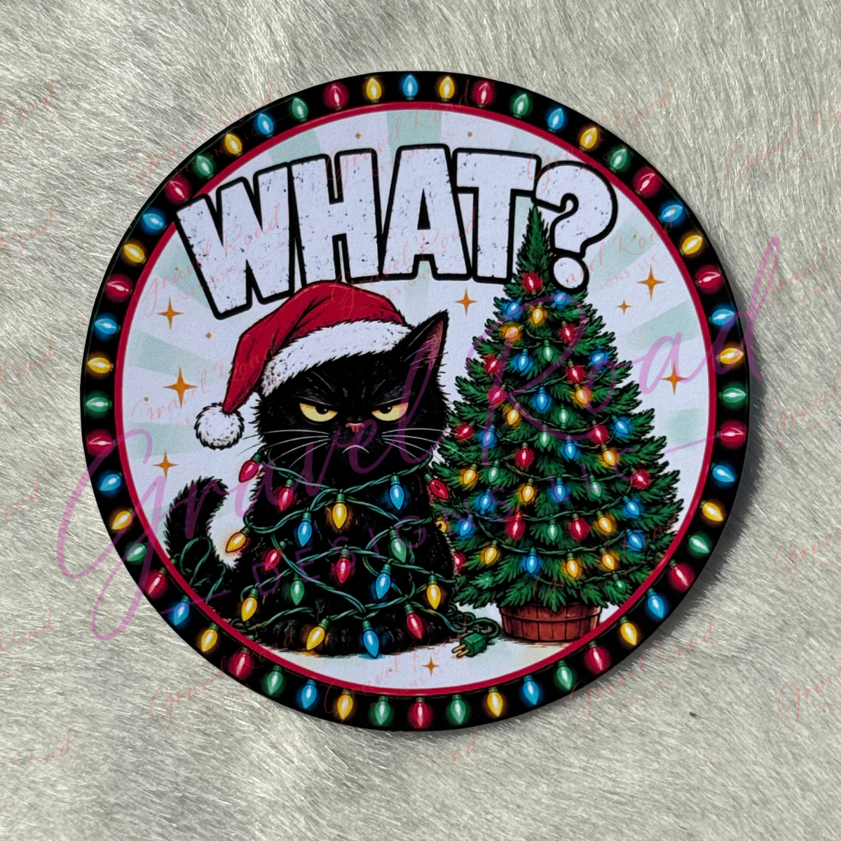 A circular cardstock featuring a black cat wearing a red Santa hat and wrapped in multicolored Christmas lights next to a decorated Christmas tree, with the word 'WHAT?' in large white letters at the top. The item is photographed on a textured gray fabric background.