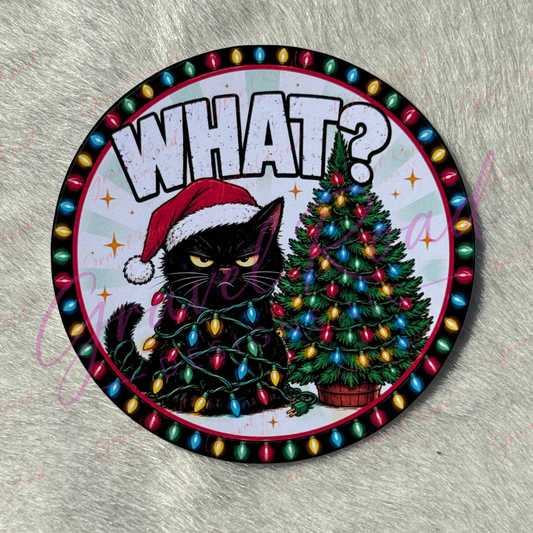 A circular cardstock featuring a black cat wearing a red Santa hat and wrapped in multicolored Christmas lights next to a decorated Christmas tree, with the word 'WHAT?' in large white letters at the top. The item is photographed on a textured gray fabric background.
