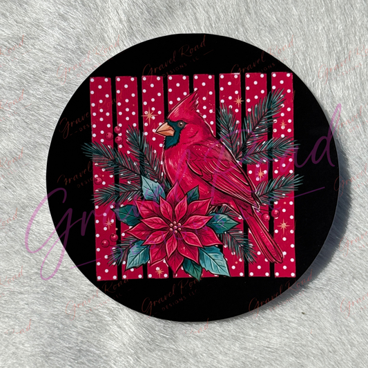A round cardstock featuring a festive holiday design with a red cardinal, red poinsettia, green pine branches, and a red-and-white polka dot stripes, photographed on a light gray textured surface.