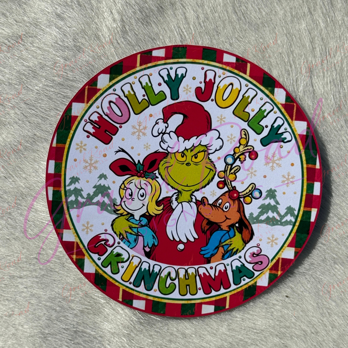 A round Christmas cardstock featuring the characters 'Holly Jolly Grinchmas' in festive red, green, yellow, and white holiday colors, photographed on a gray faux-fur surface.