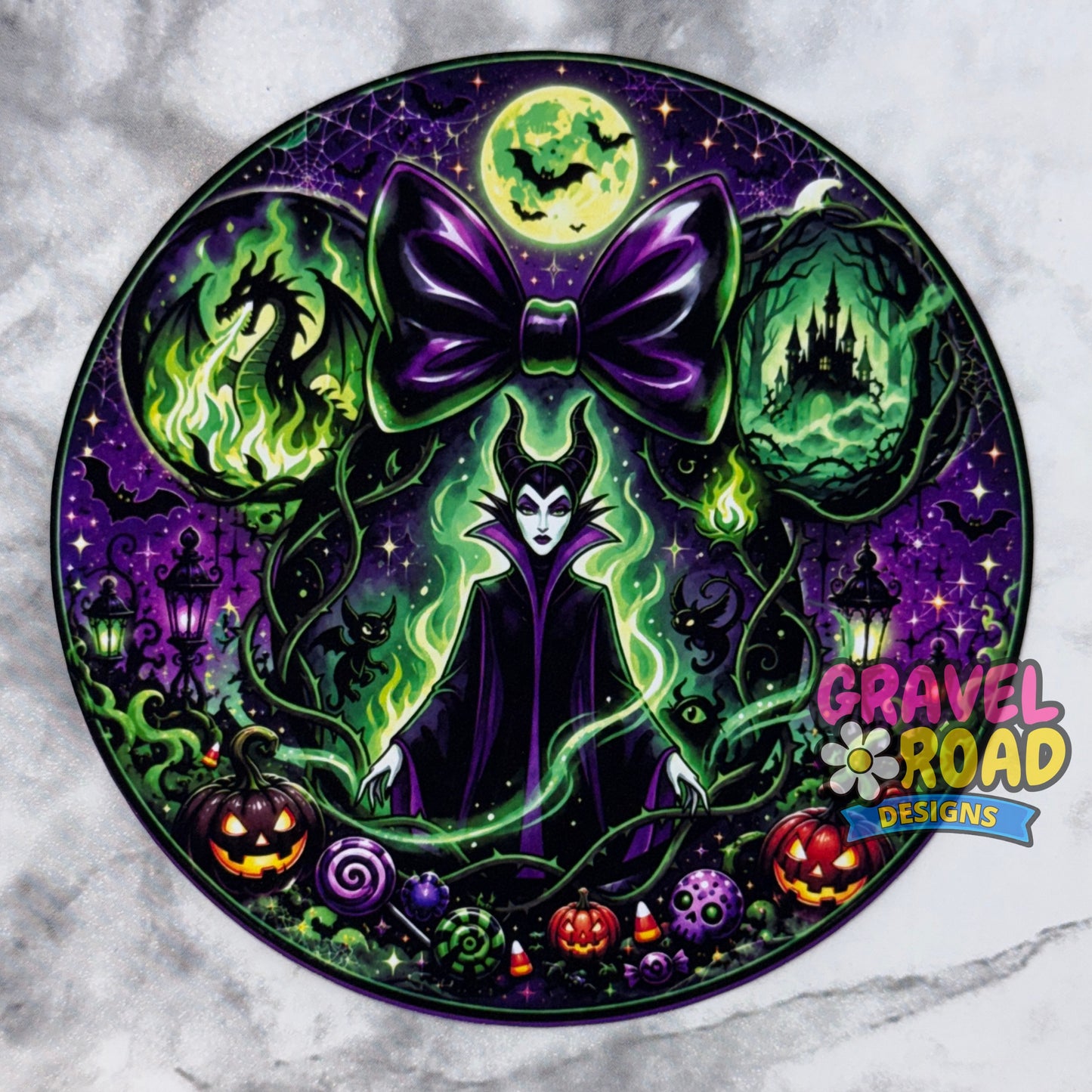 A circular Halloween-themed cardstock featuring a central female witch character with a large purple bow, surrounded by green magical vines, jack-o'-lanterns, candy canes, and haunted house imagery on a purple night sky. The design includes a 'Gravel Road Designs' logo in the bottom right corner. On a grey marble backdrop.