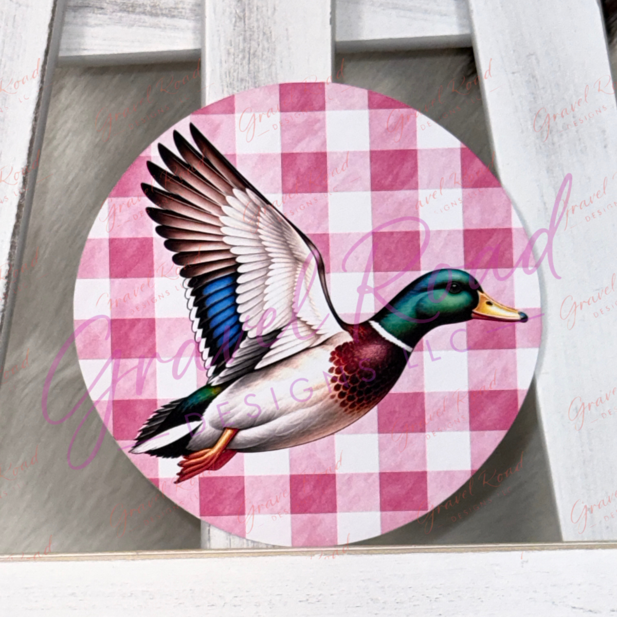 A circular cardstock featuring a detailed illustration of a mallard duck in flight over a pink and white checkered background, displayed on a rustic white wooden surface.