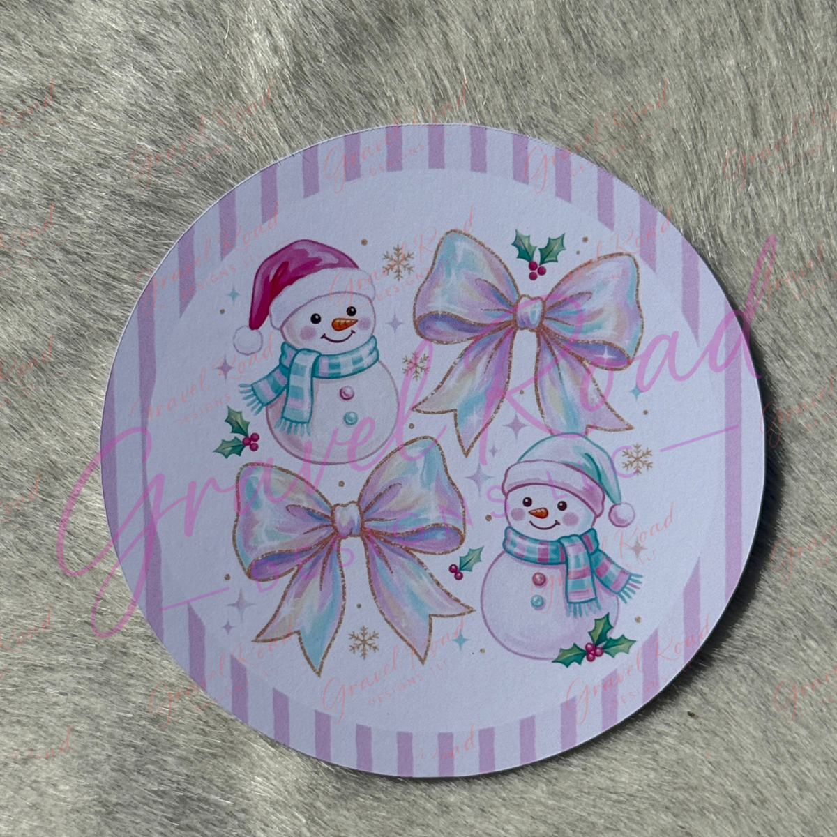 A round printed cardstock featuring a Christmas design with two snowmen, two large iridescent bows, and small gold snowflakes on a lavender striped border, placed on a gray furry surface.