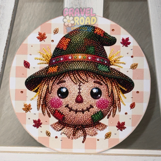 Round fall cardstock featuring a cute patchwork scarecrow with a brown hat, rosy cheeks, and scattered autumn leaves. The scarecrow has the look of rhinestones.