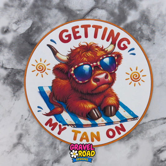 Cardstock with a cartoon highland cow wearing sunglasses on a towel, text 'Getting My Tan On', and Gravel Road Designs logo on a marble background.