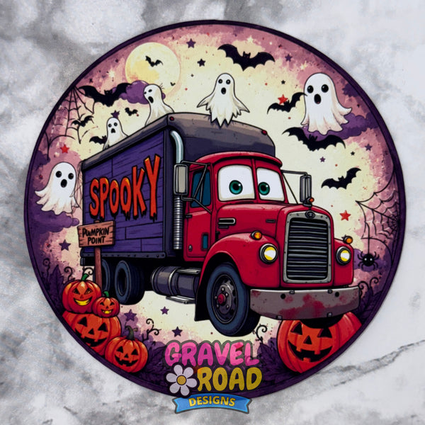 A circular Halloween-themed cardstock featuring a red cartoon truck with eyes and a grille, labeled 'Spooky' on its box, surrounded by ghosts, bats, pumpkins, and a purple spiderweb background. The design includes the logo 'Gravel Road Designs' at the bottom. On a grey marble backdrop.
