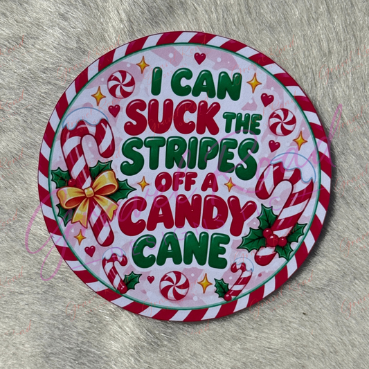 A round Christmas-themed cardstock with the text 'I CAN SUCK THE STRIPES OFF A CANDY CANE' in green and red lettering. The design features red and white striped candy canes, green holly leaves, red hearts, and yellow stars on a light pink background, surrounded by a red and white striped border. Displayed on a faux fur background.