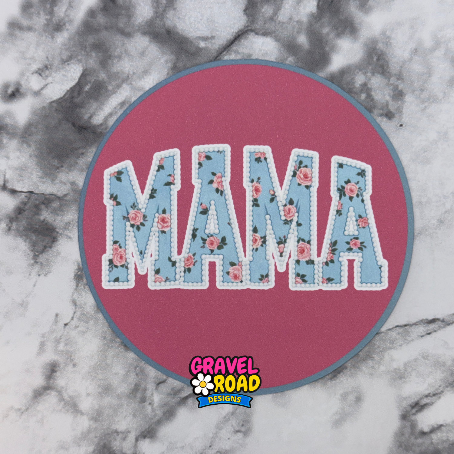 Circular cardstock printed in pink with the word "Mama" in blue with flowers. Gravel Road Designs logo printed on the  bottom.