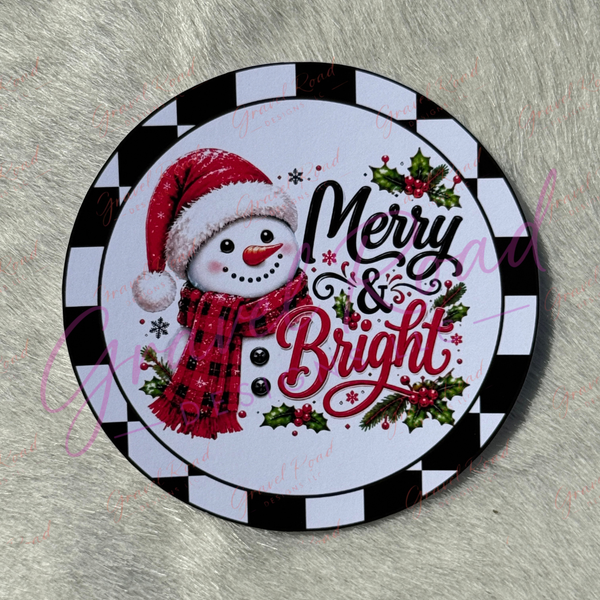 A round holiday-themed cardstock featuring a snowman in a red Santa hat and red plaid scarf beside the words “Merry & Bright,” with holly and a black-and-white checkered border, shown on a gray faux-fur surface.