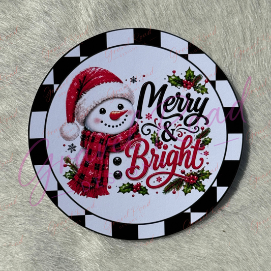 A round holiday-themed cardstock featuring a snowman in a red Santa hat and red plaid scarf beside the words “Merry & Bright,” with holly and a black-and-white checkered border, shown on a gray faux-fur surface.