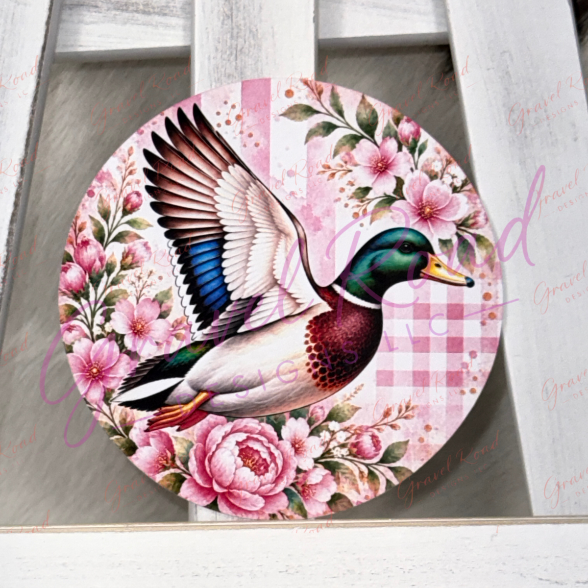 A round cardstock featuring a detailed illustration of a mallard duck in flight surrounded by pink flowers and a pink gingham pattern, displayed on a white wooden surface.