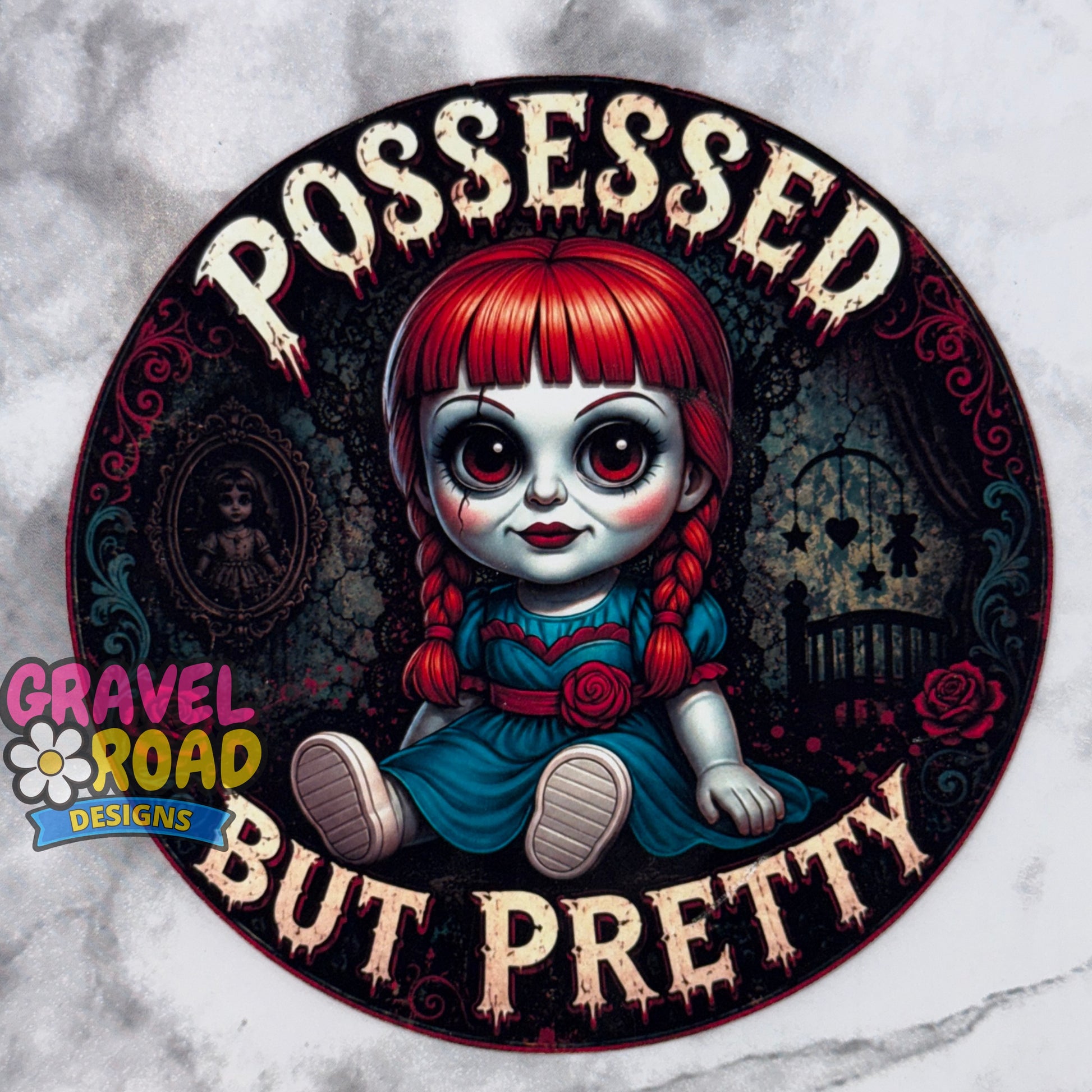 A circular cardstock design featuring a creepy doll character with red hair, large eyes, and a blue dress, surrounded by the text 'POSSESSED BUT PRETTY' in a dripping font. A 'Gravel Road Designs' logo is visible on the bottom left. On a grey marble backdrop.