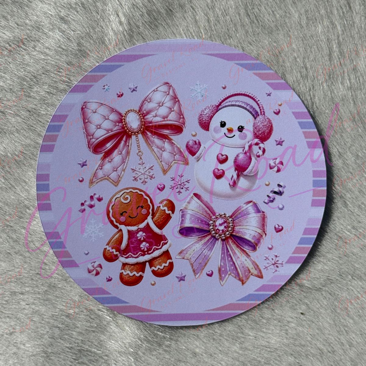 A round printed cardstock featuring a festive Christmas design with a snowman, gingerbread cookie, and two large pink bows on a lavender background, displayed on a gray faux fur surface.