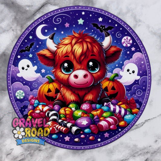 A circular Halloween-themed cardstock featuring a cute orange-brown fluffy cow surrounded by candy, pumpkins, ghosts, stars, and bats on a purple night sky. The design includes the logo 'GRAVEL ROAD DESIGNS' in the lower left and is shown on a light marble-like background.