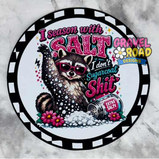 A circular cardstock design featuring a raccoon wearing pink sunglasses and a salt shaker labeled 'Extra Salty,' with the text 'I season with SALT I don't sugarcoat shit.' The design includes the brand/logo 'Gravel Road Designs' and is shown on a light gray marble-like background.