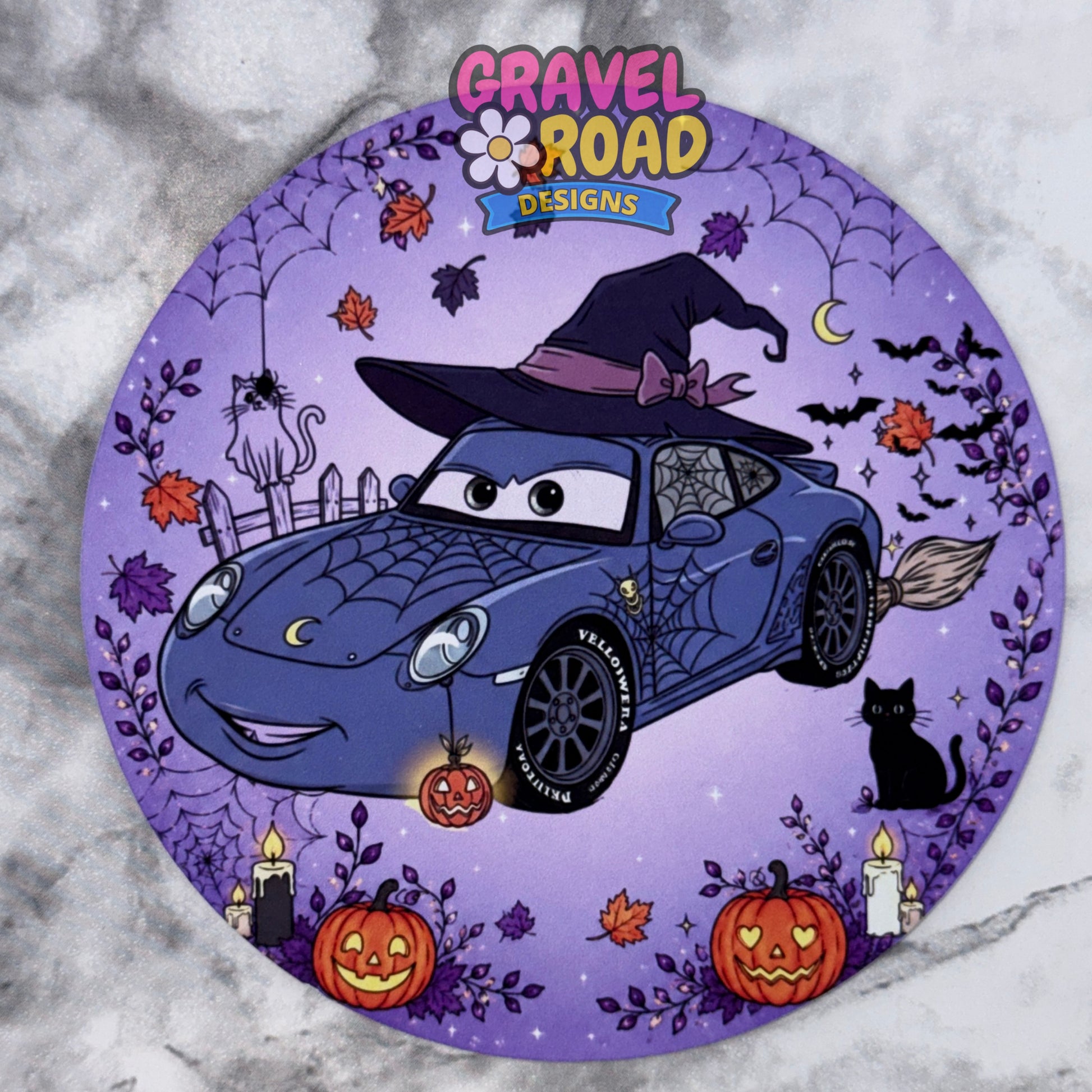 A circular Halloween-themed cardstock featuring a purple car with spiderwebs, a witch hat, jack-o'-lanterns, candles, bats, and a cat on a purple background, with the logo 'Gravel Road Designs' at the top. On a grey marble backdrop.