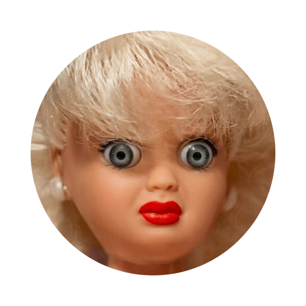 A close-up circular photo of a vintage blonde doll or collectible figure with large gray-blue eyes, red lips, and light pink skin, isolated on a black background.