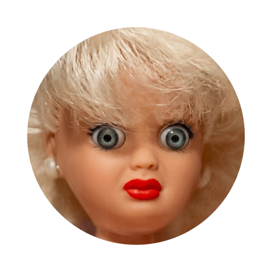 A close-up circular photo of a vintage blonde doll or collectible figure with large gray-blue eyes, red lips, and light pink skin, isolated on a black background.
