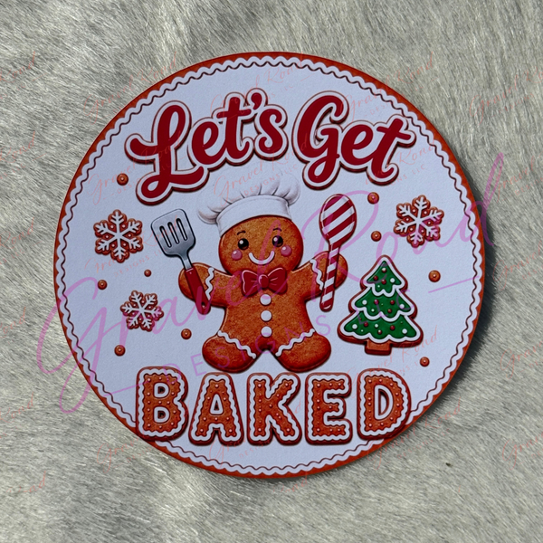 A round baking-themed cardstock featuring a gingerbread person in a chef hat with the words 'Let's Get BAKED,' surrounded by red snowflakes, a green Christmas tree, and a spatula, displayed on a gray furry surface.