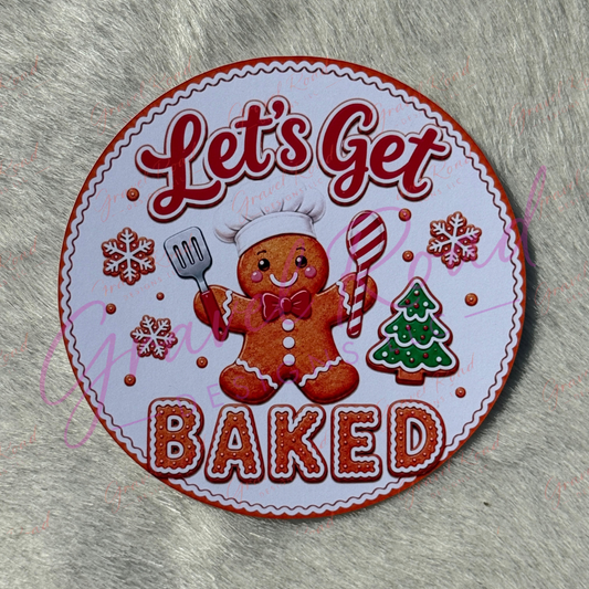 A round baking-themed cardstock featuring a gingerbread person in a chef hat with the words 'Let's Get BAKED,' surrounded by red snowflakes, a green Christmas tree, and a spatula, displayed on a gray furry surface.