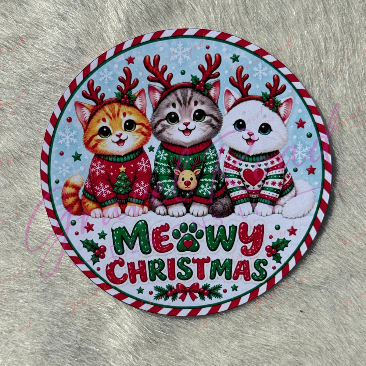 A round Christmas-themed cardstock featuring three cute cartoon cats wearing holiday sweaters and reindeer antlers, with the text 'MEOWY CHRISTMAS' at the bottom, photographed on a gray faux fur surface.