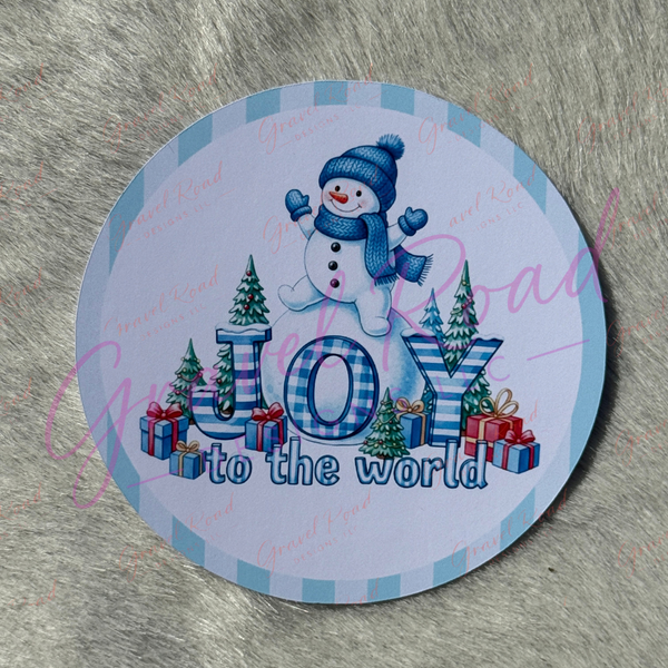 A round cardstock featuring a winter holiday design with a snowman, the text 'JOY to the world,' Christmas trees, and wrapped gifts on a light blue and white plaid border, placed on a gray faux fur surface.