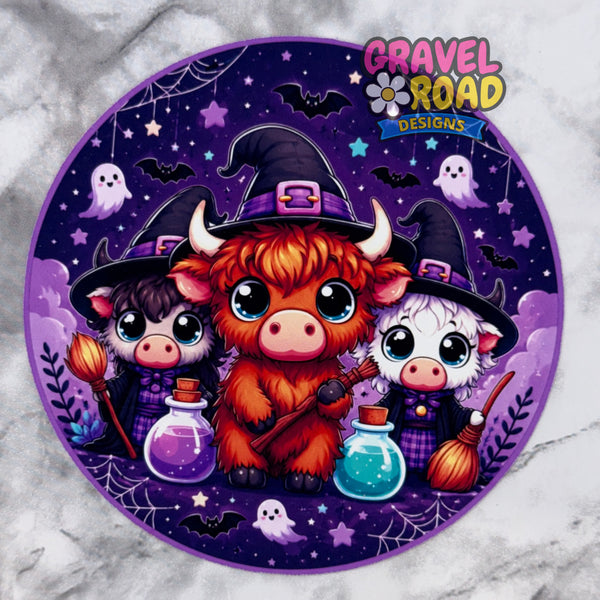A circular Halloween-themed cardstock design featuring three cute cartoon animals dressed as witches, with purple hats, broomsticks, potion bottles, ghosts, bats, and spiderwebs on a dark purple background. The logo 'Gravel Road Designs' appears in the top right corner.