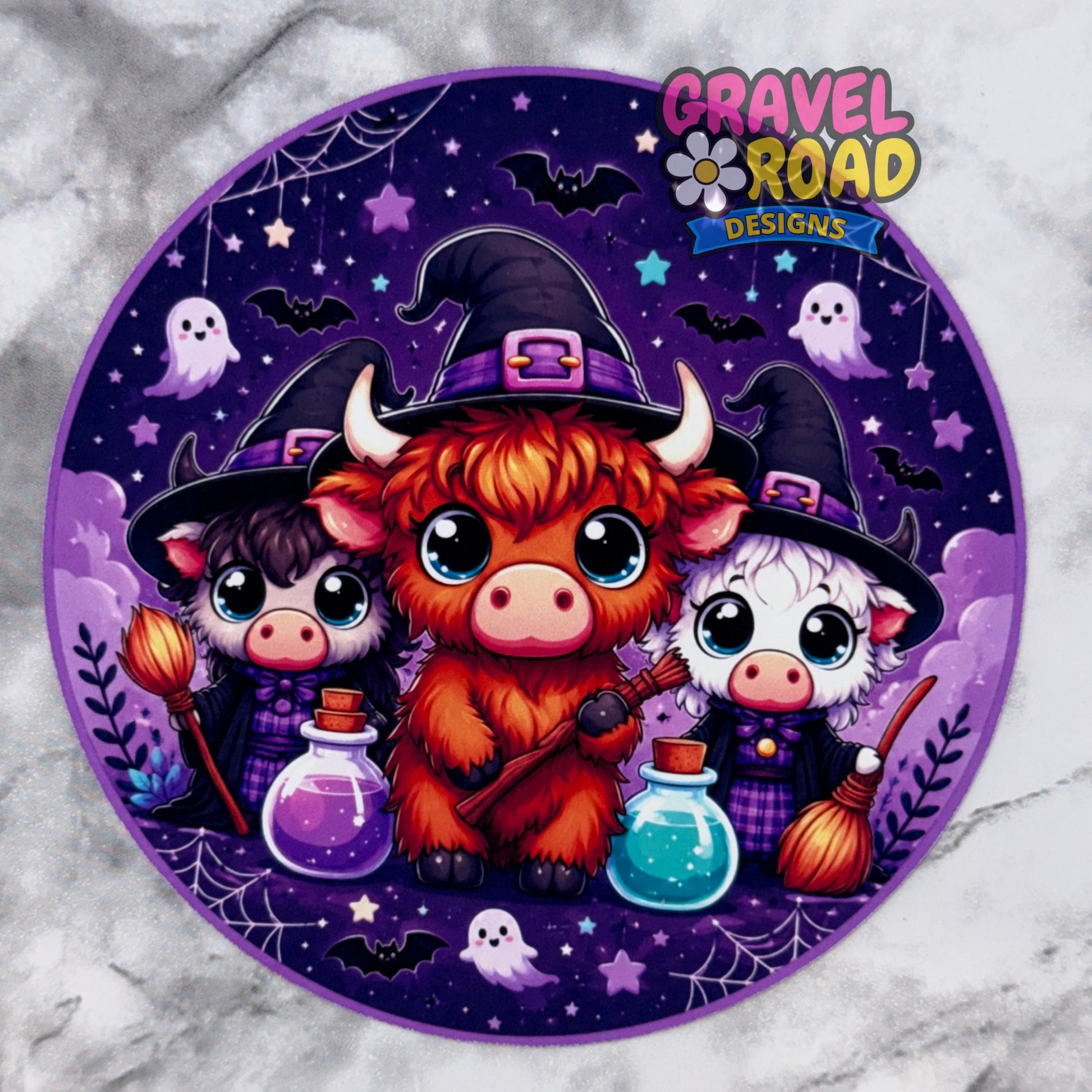 A circular Halloween-themed cardstock design featuring three cute cartoon animals dressed as witches, with purple hats, broomsticks, potion bottles, ghosts, bats, and spiderwebs on a dark purple background. The logo 'Gravel Road Designs' appears in the top right corner.