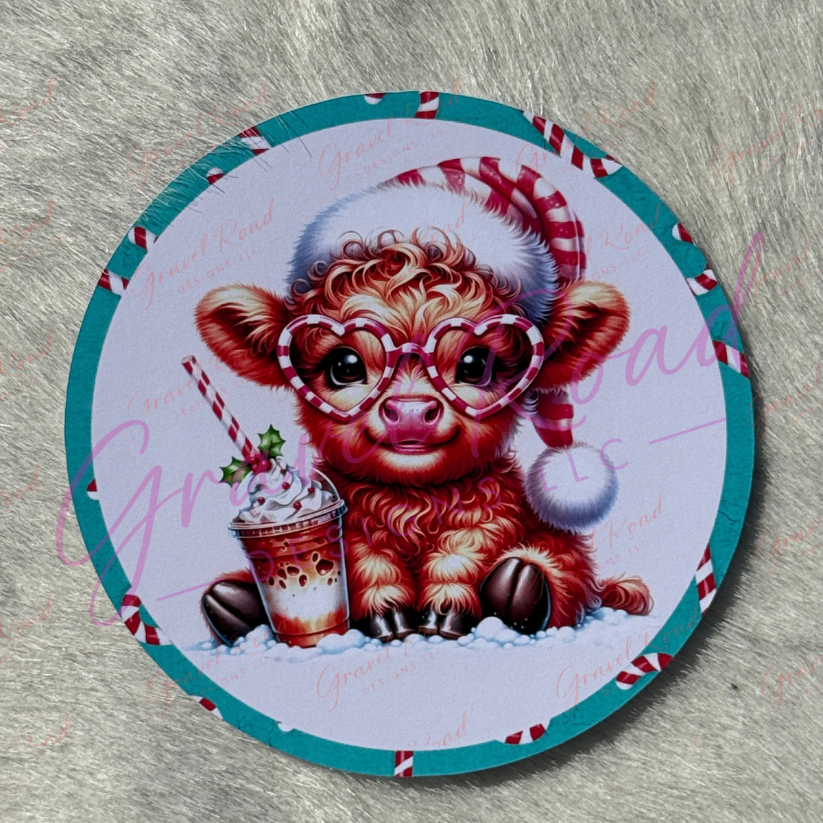 A round printed cardstock featuring a cute cartoon Highland Cow wearing a Santa hat and heart-shaped glasses, sitting in snow and holding a drink, with a teal border and red-and-white candy cane accents. The item is photographed on a gray textured surface with a faint watermark overlay.