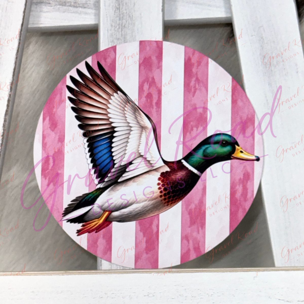A round cardstock featuring a flying mallard duck over pink and white vertical stripes, displayed on a white wooden surface.