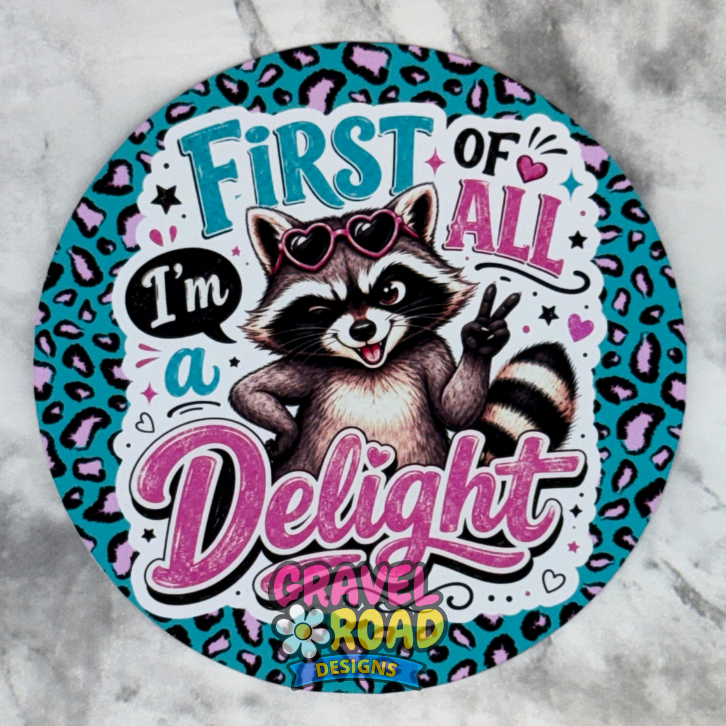 A circular printed cardstock design featuring a cartoon raccoon wearing heart-shaped sunglasses and making a peace sign, surrounded by the text 'FIRST OF ALL I'm a Delight" with the logo "Gravel Road Designs" on a turquoise leopard-print background, shown on a light marble surface.