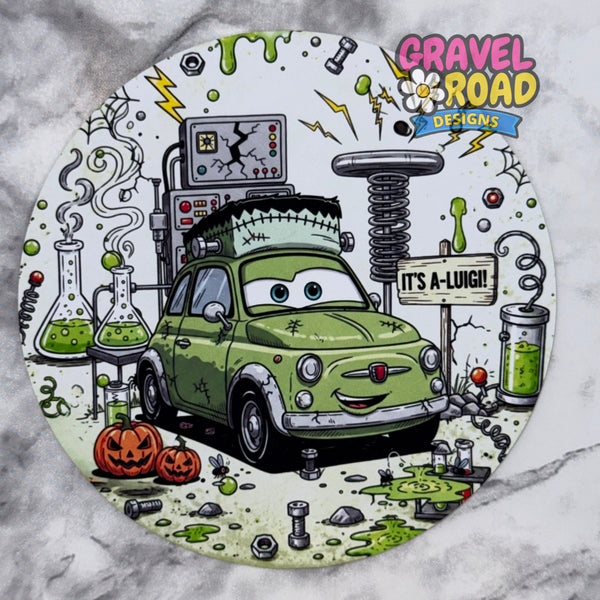 A circular cardstock featuring a cartoon green car styled like Frankenstein's monster, surrounded by Halloween-themed elements including pumpkins, test tubes with green liquid, a spring machine, and the text 'IT'S A-LUIGI!' and the logo 'Gravel Road Designs'. On a grey marble backdrop.