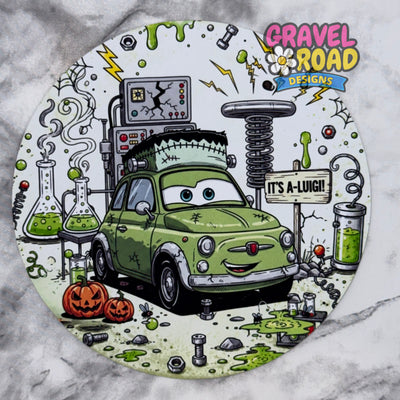 A circular cardstock featuring a cartoon green car styled like Frankenstein's monster, surrounded by Halloween-themed elements including pumpkins, test tubes with green liquid, a spring machine, and the text 'IT'S A-LUIGI!' and the logo 'Gravel Road Designs'. On a grey marble backdrop.