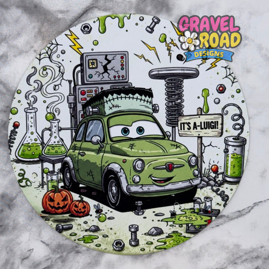 A circular cardstock featuring a cartoon green car styled like Frankenstein's monster, surrounded by Halloween-themed elements including pumpkins, test tubes with green liquid, a spring machine, and the text 'IT'S A-LUIGI!' and the logo 'Gravel Road Designs'. On a grey marble backdrop.
