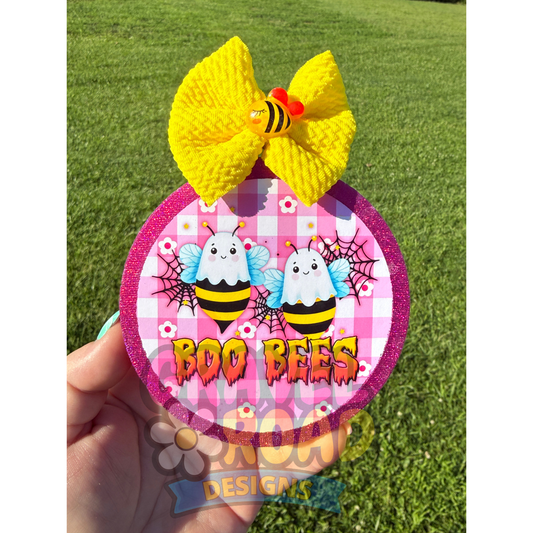 A hand holds a circular Halloween-themed accessory featuring two cartoon bees on a pink gingham background with the words 'BOO BEES' and a large yellow knitted bow on top. The background is an outdoor grassy lawn.