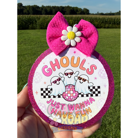 A hand holds a round car freshie outdoors over a grassy field. The item has a bright pink glitter border, a large pink bow on top with a white and yellow flower center, and a printed design featuring two cartoon ghosts, sunglasses, a pink disco ball, and the text 'GHOULS JUST WANNA HAVE FUN'.