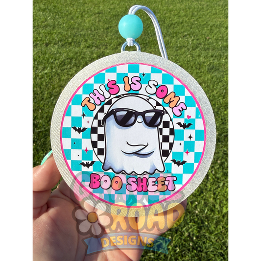 A hand holds a round glittery Halloween-themed car freshie against a green grass background. The sheet features a cartoon ghost wearing black sunglasses, a black-and-white checkered headband, and a turquoise and white checkered background with the text 'THIS IS SOME BOO SHEET' in pink and orange lettering.