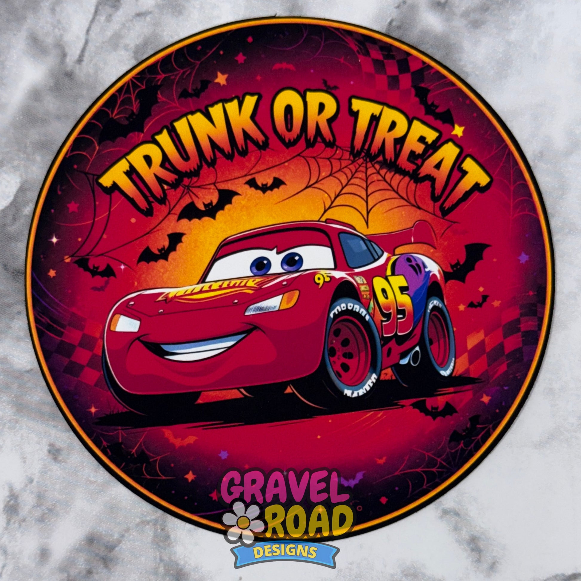 A circular Halloween-themed cardstock featuring a race car with the text 'TRUNK OR TREAT' on a red, orange, and purple spooky background with bats and spiderwebs. Gravel Road Designs logo. On a grey marble background.