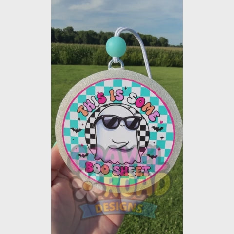 A hand holds a round glittery Halloween-themed car freshie against a green grass background. The sheet features a cartoon ghost wearing black sunglasses, a black-and-white checkered headband, and a turquoise and white checkered background with the text 'THIS IS SOME BOO SHEET' in pink and orange lettering.