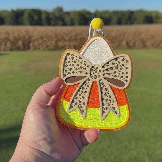 A video shows a hand holding a candy corn-shaped car freshie outdoors against a blurred field and sky background. The car freshie has a beige bow with dark spots, orange and yellow glitter sections, and a silver hanging loop at the top.