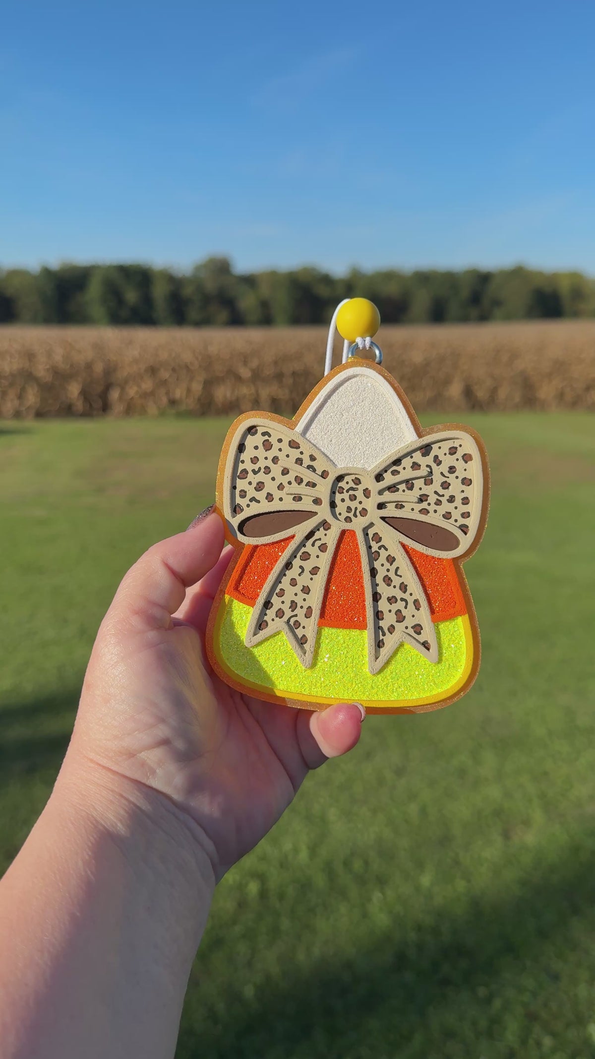 A video shows a hand holding a candy corn-shaped car freshie outdoors against a blurred field and sky background. The car freshie has a beige bow with dark spots, orange and yellow glitter sections, and a silver hanging loop at the top.