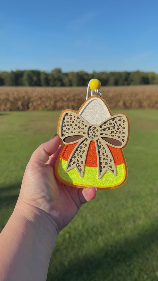 A video shows a hand holding a candy corn-shaped car freshie outdoors against a blurred field and sky background. The car freshie has a beige bow with dark spots, orange and yellow glitter sections, and a silver hanging loop at the top.