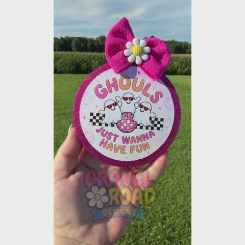 A hand holds a round car freshie outdoors over a grassy field. The item has a bright pink glitter border, a large pink bow on top with a white and yellow flower center, and a printed design featuring two cartoon ghosts, sunglasses, a pink disco ball, and the text 'GHOULS JUST WANNA HAVE FUN'.